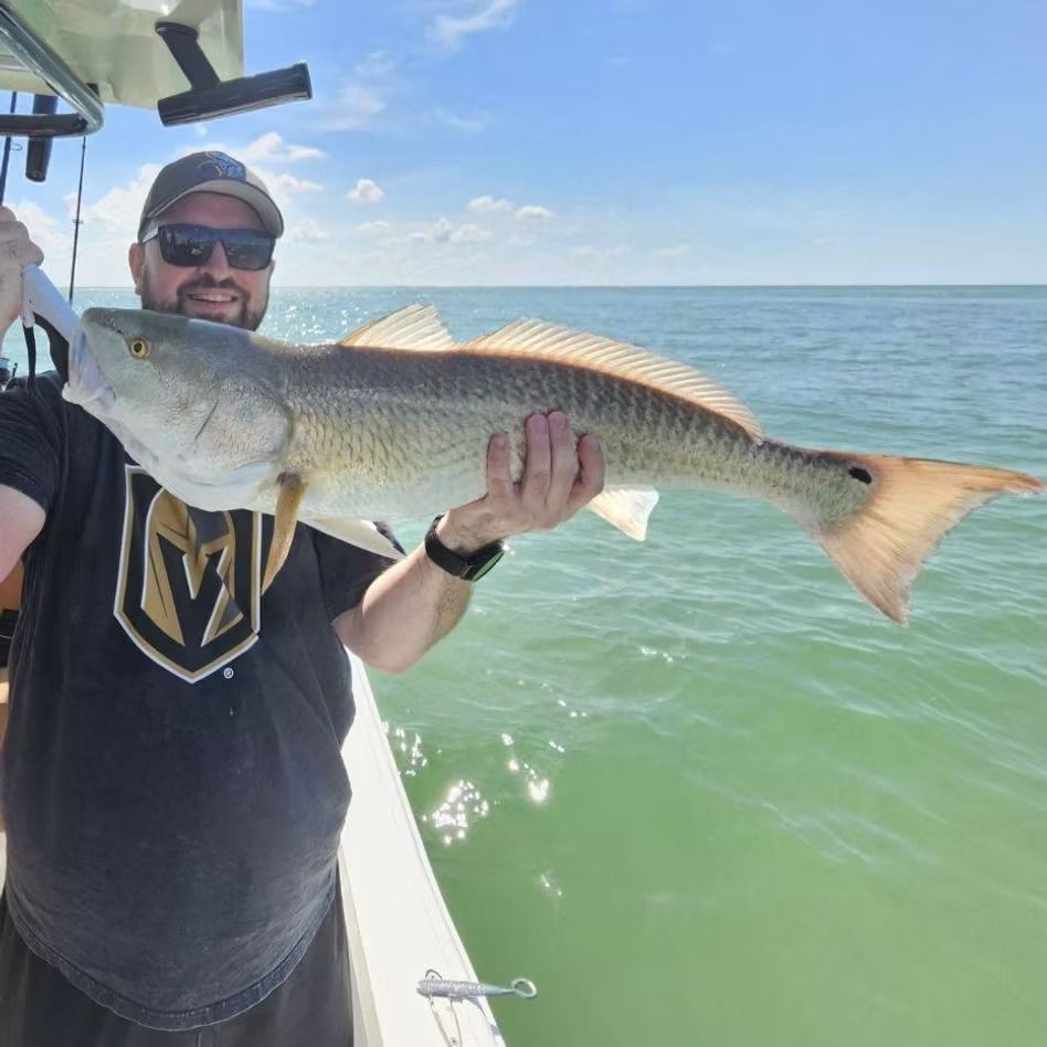Man holding a large redfish on a boat, smiling. Sunny day, ocean background.