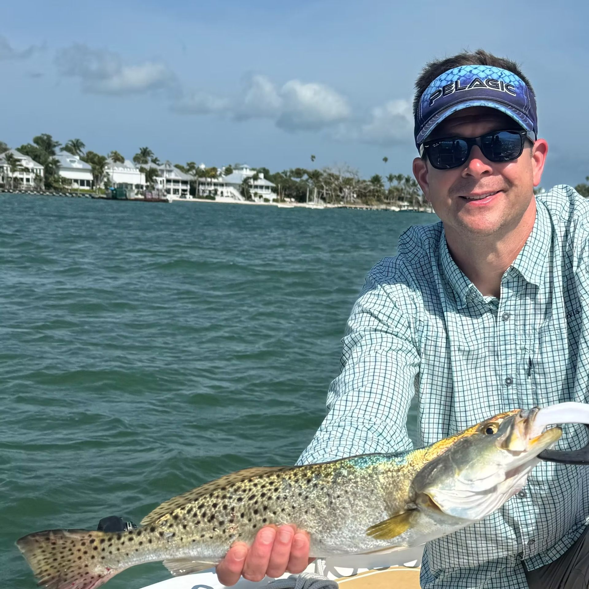 Man on boat holding a speckled trout; houses and water in the background.
