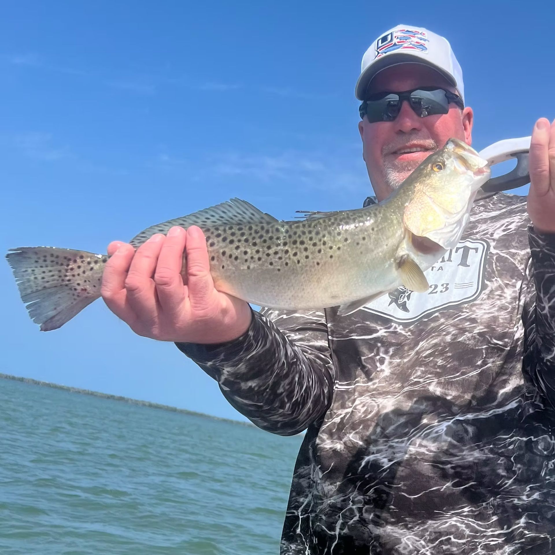 Man holding a speckled trout fish on a boat, sunny day.