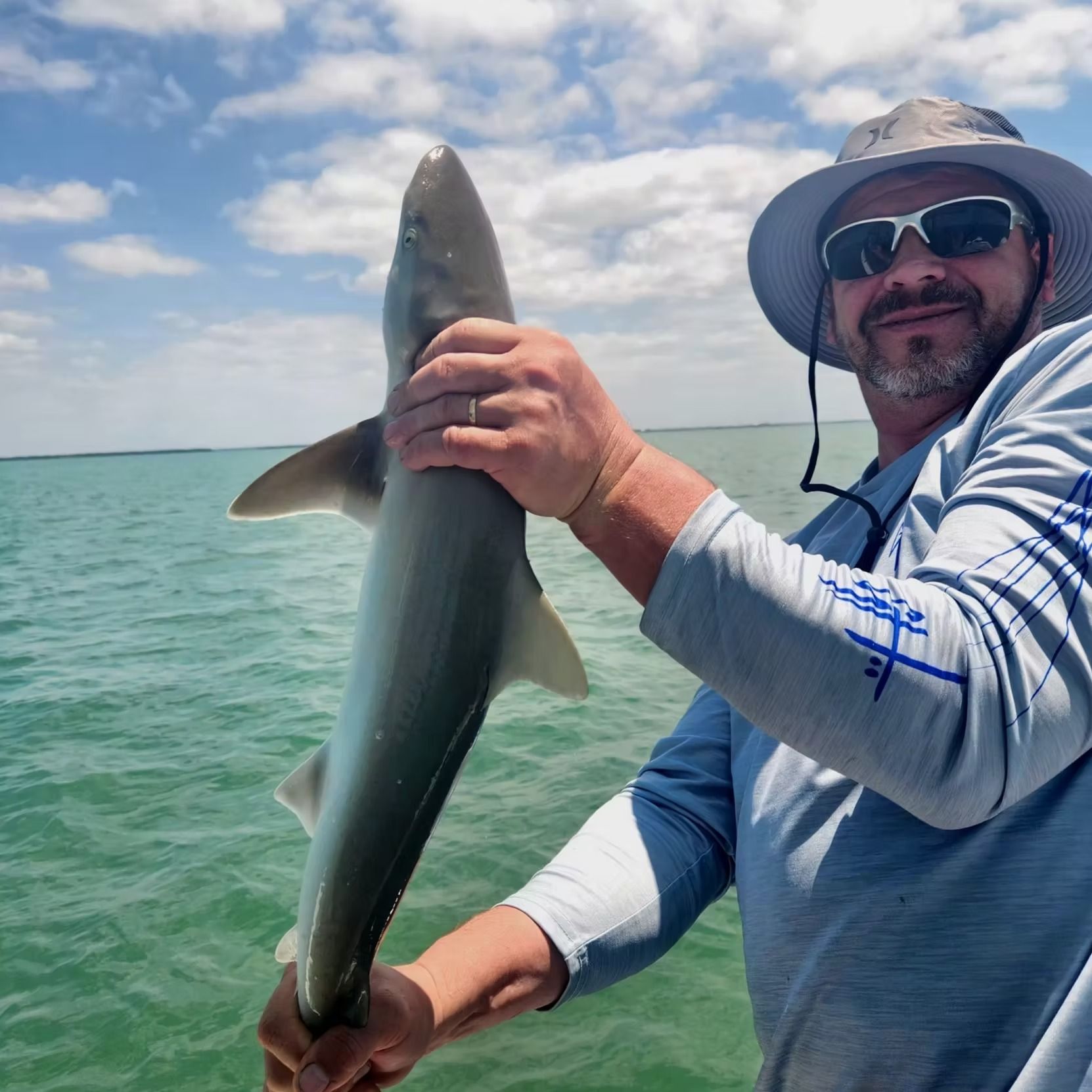 Man holding a small shark he caught, near blue ocean, under a sunny sky.