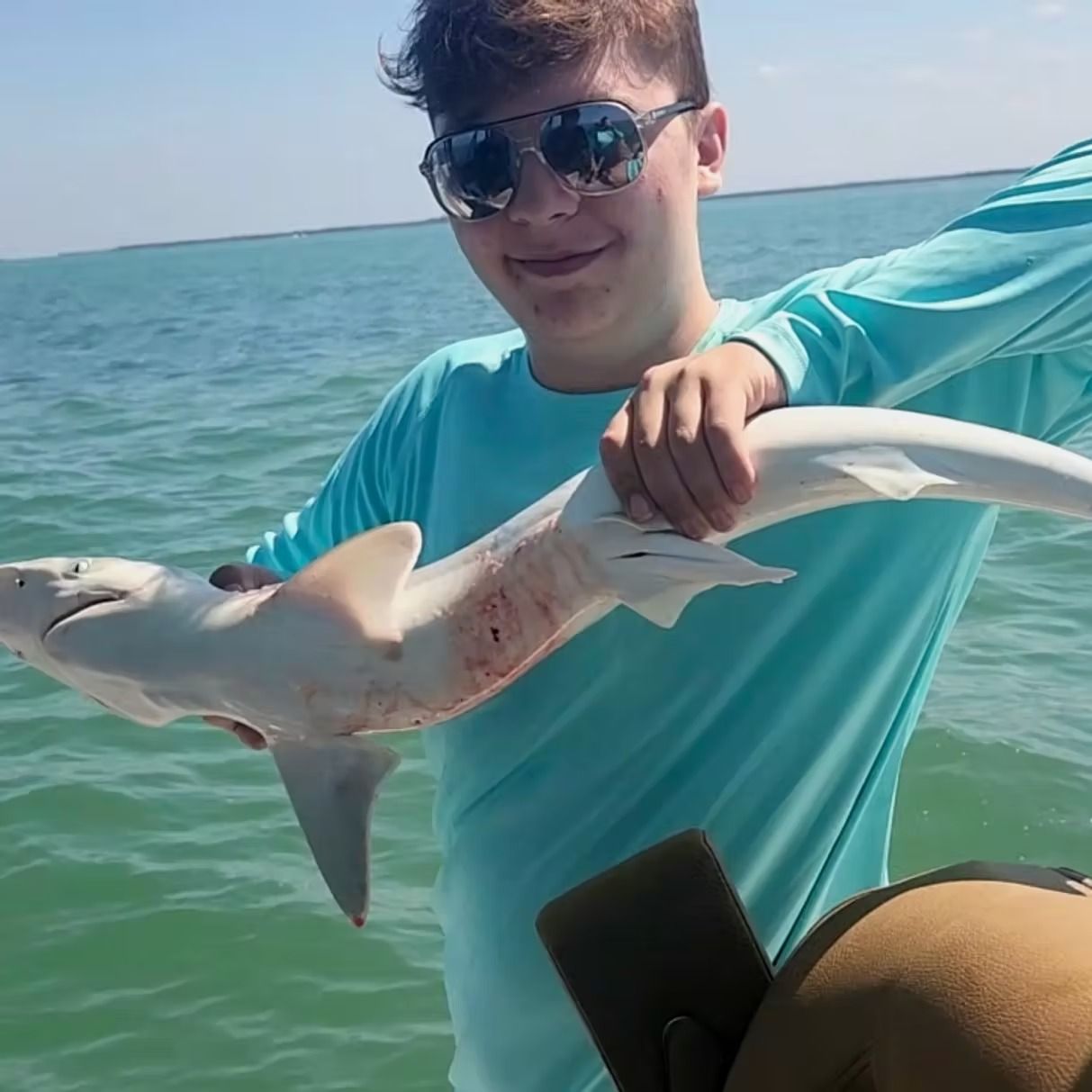 Teenager smiling, holding a small shark on a boat. Ocean in background, sunny day.