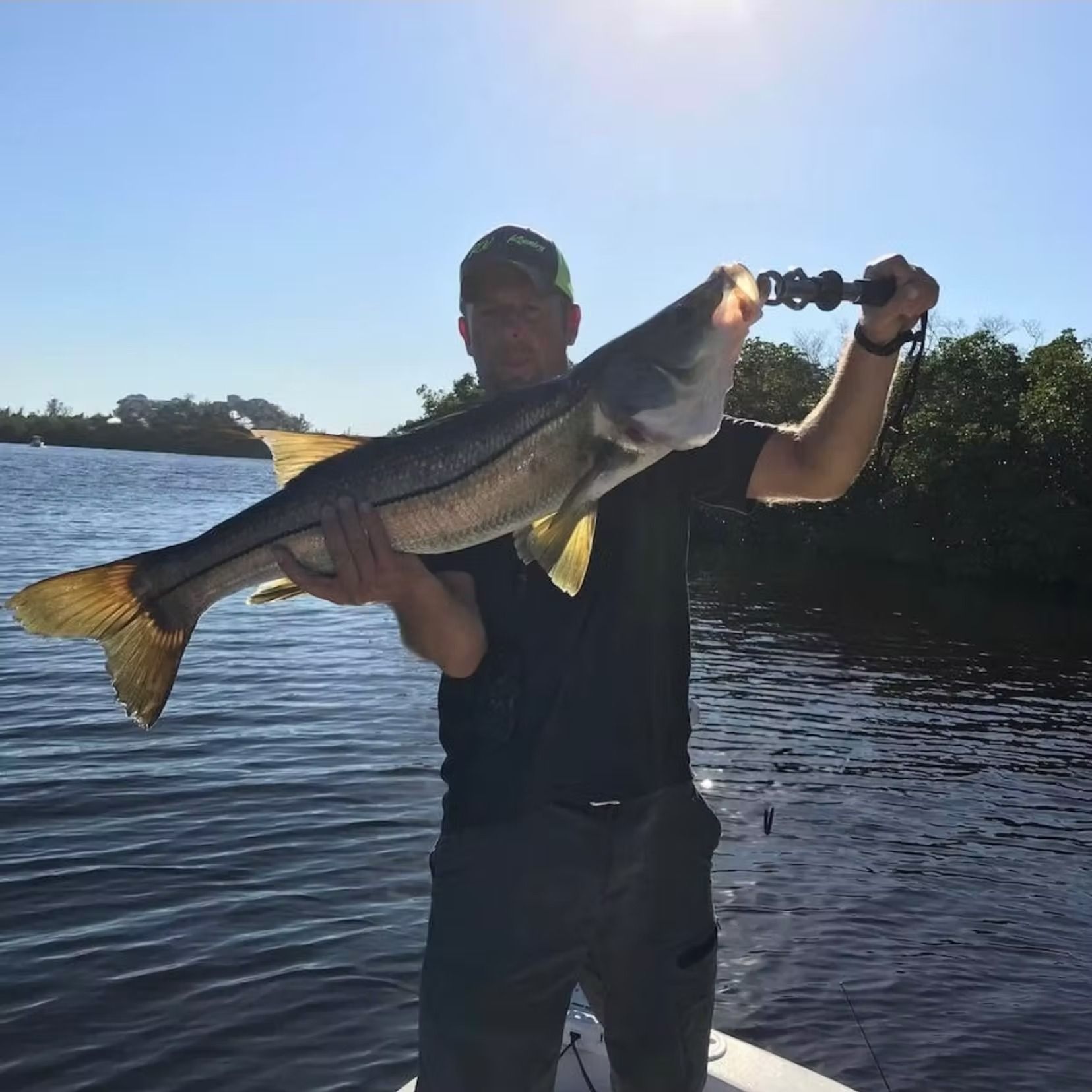 Man holding a large snook fish on a boat, outdoors on a sunny day.