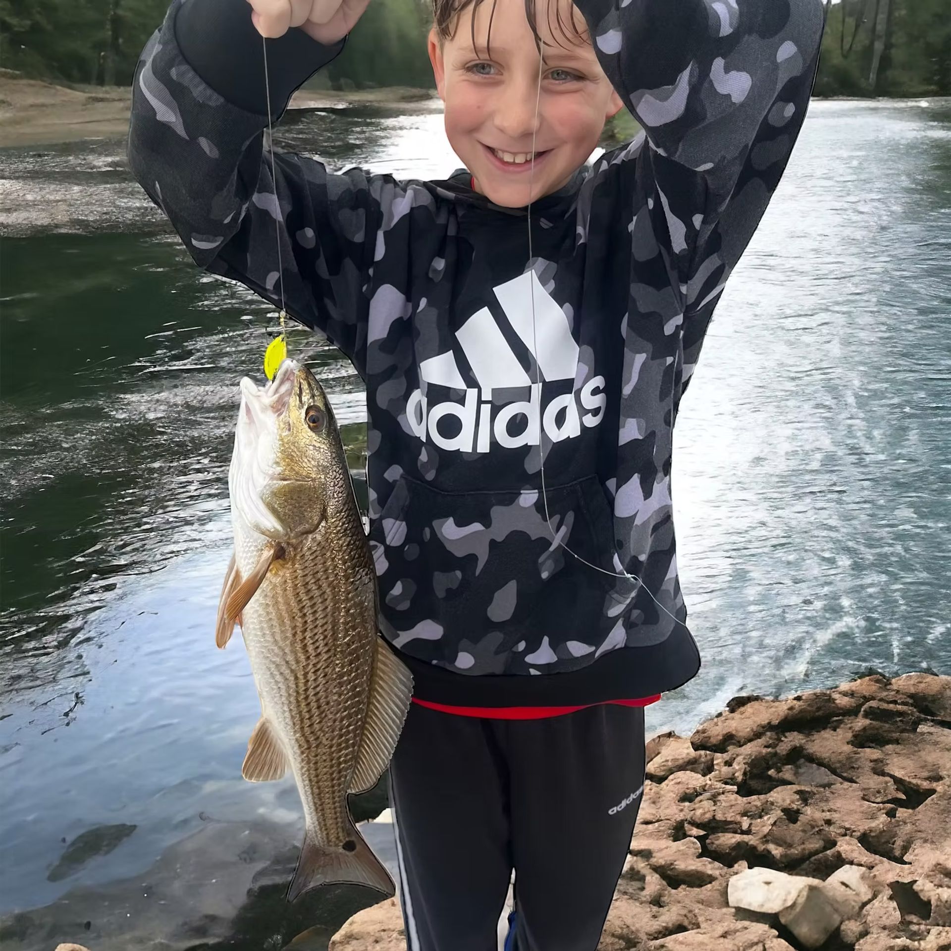 Boy in camo hoodie smiles, holding up a fish with a lure near a river.