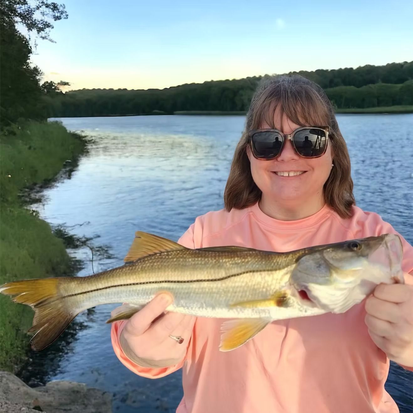 Woman smiling, holding a fish near a river, wearing sunglasses and a pink shirt.