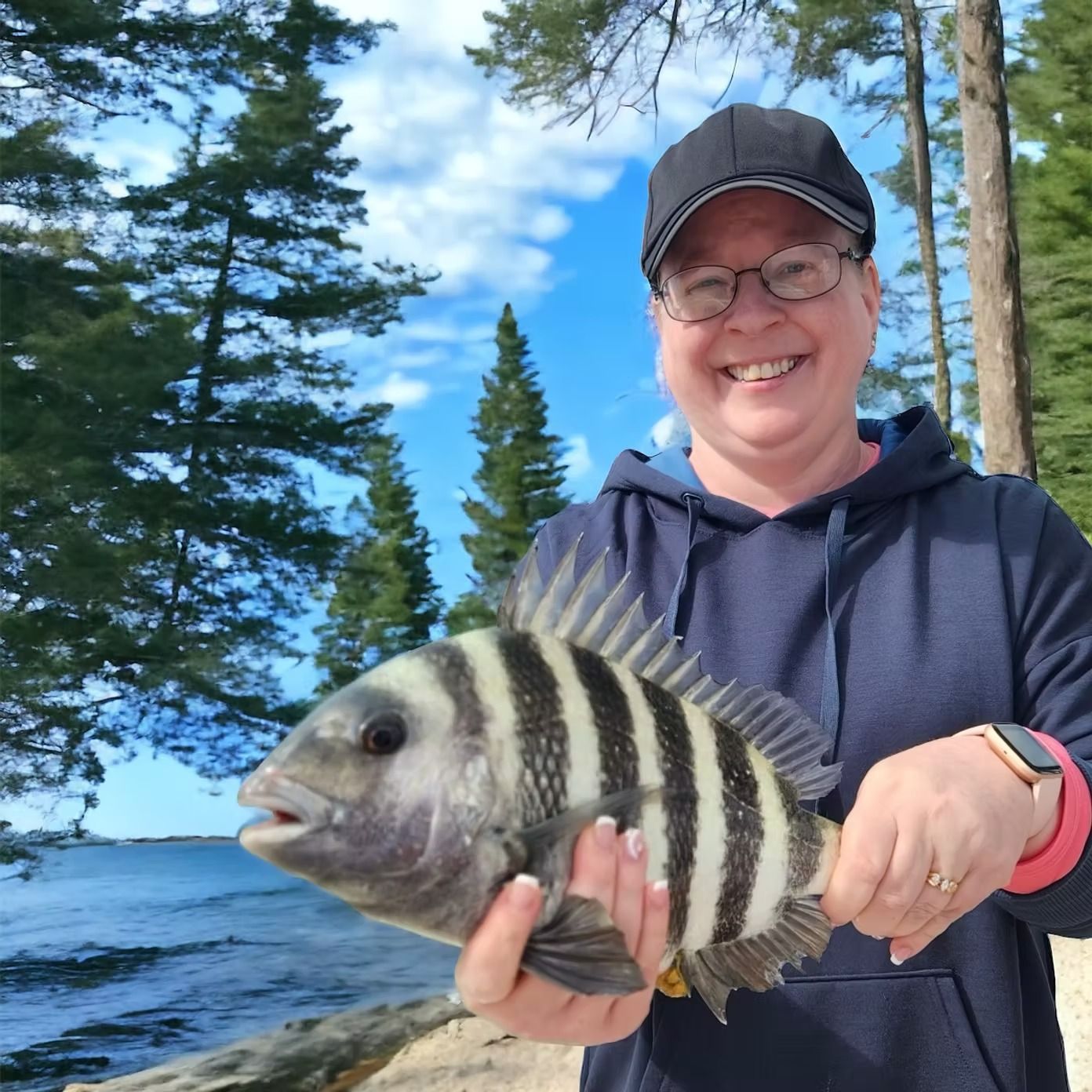 Woman holding a striped sheepshead fish by the water, smiling. Blue hoodie, black cap, outdoors.