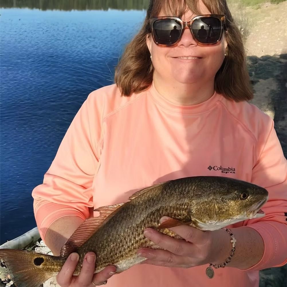 Woman in sunglasses holding a redfish near water. She wears peach long-sleeved shirt, smiles.