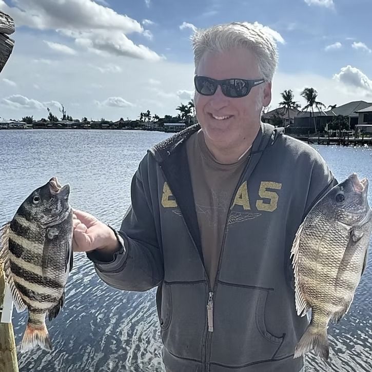 Man holding two fish he caught by the water, wearing sunglasses and a hoodie, smiling.
