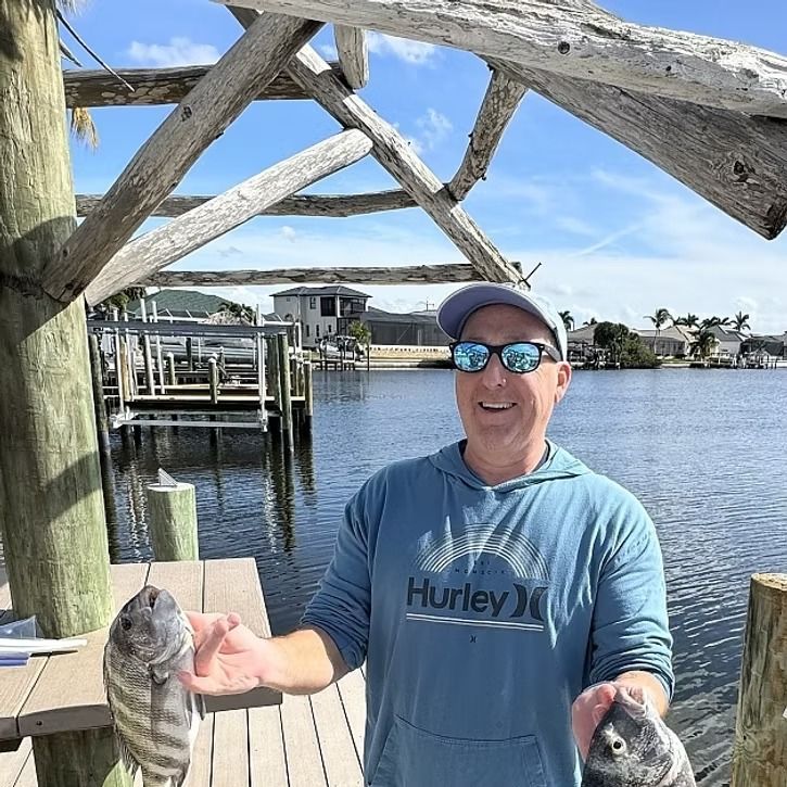 Man fishing on a wooden dock, holding two fish. Blue sky, water, and wearing a Hurley shirt and sunglasses.