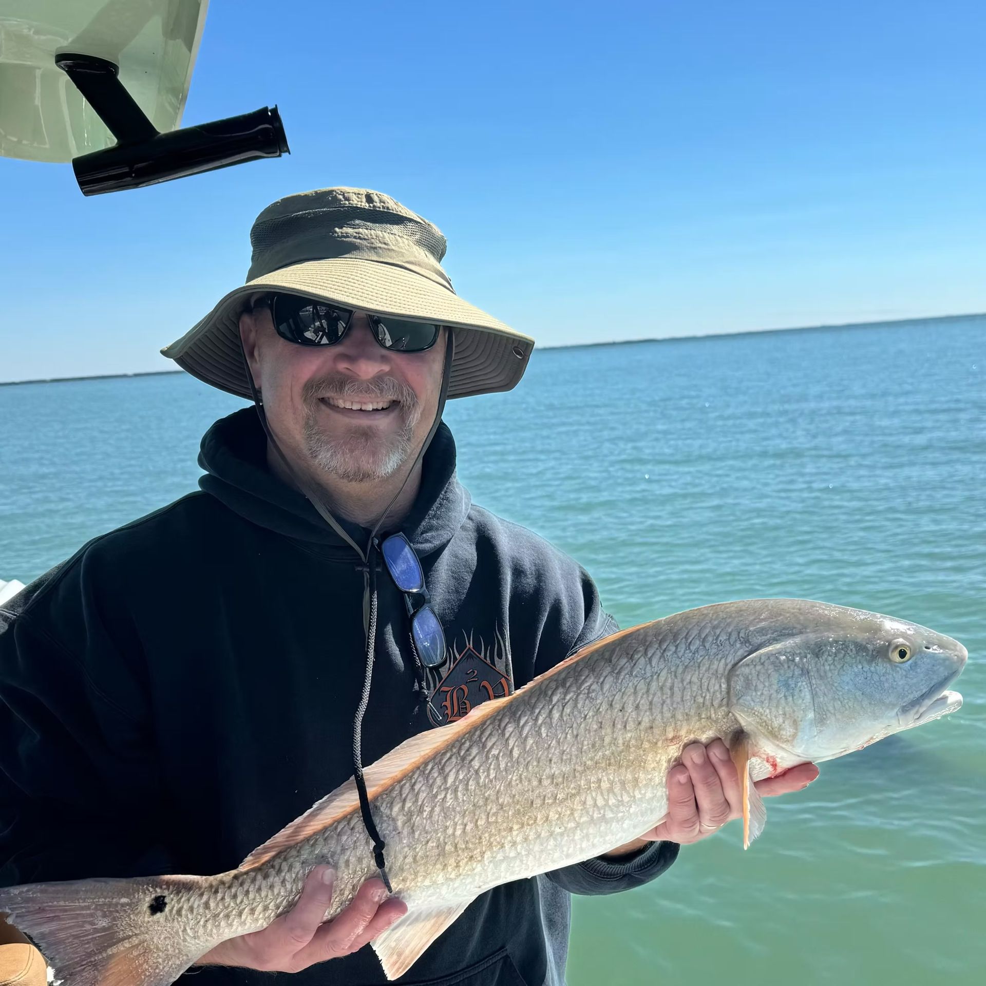 Man wearing hat and sunglasses holding a large reddish fish on a boat, ocean in background.