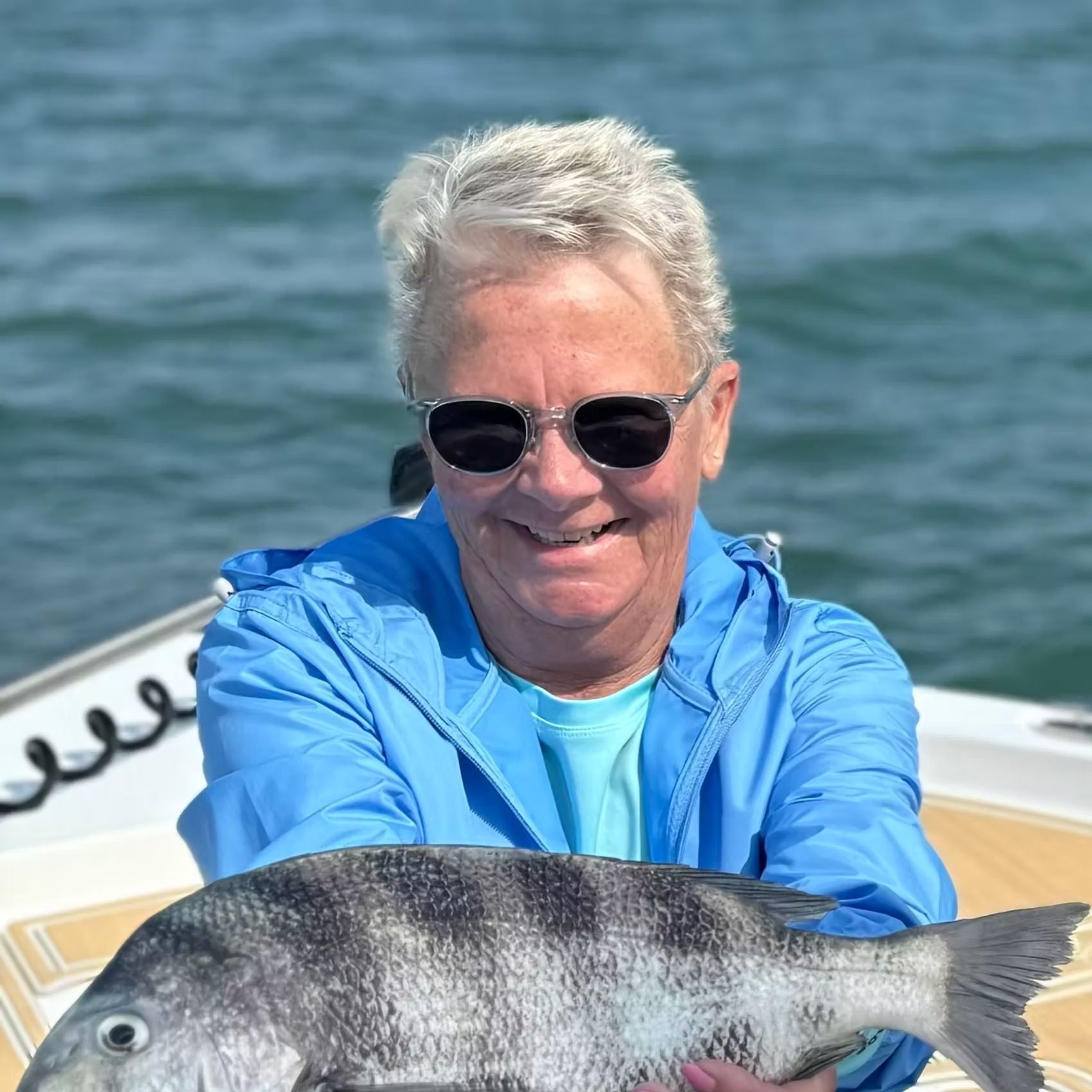 Woman on a boat holding a fish, smiling, wearing sunglasses and blue jacket, water in the background.