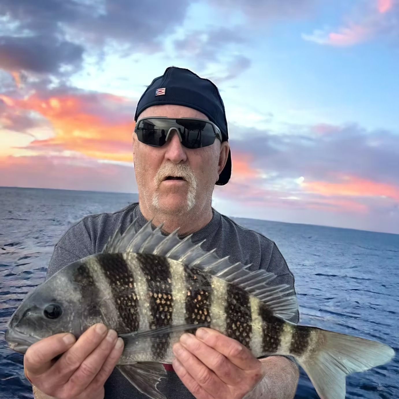 Man holding a striped fish at sea with a sunset in the background.