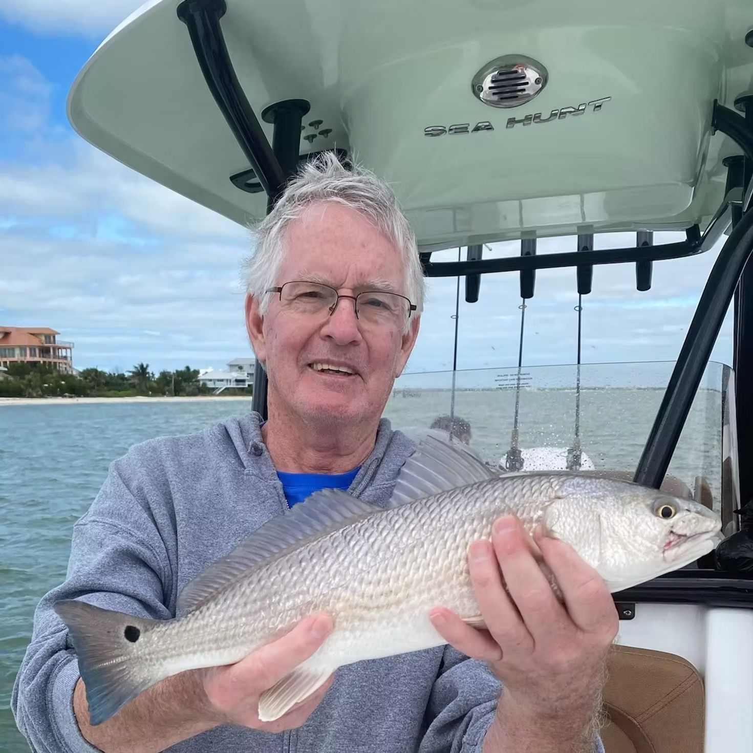Man holding a redfish on a boat; water and coastline in background.