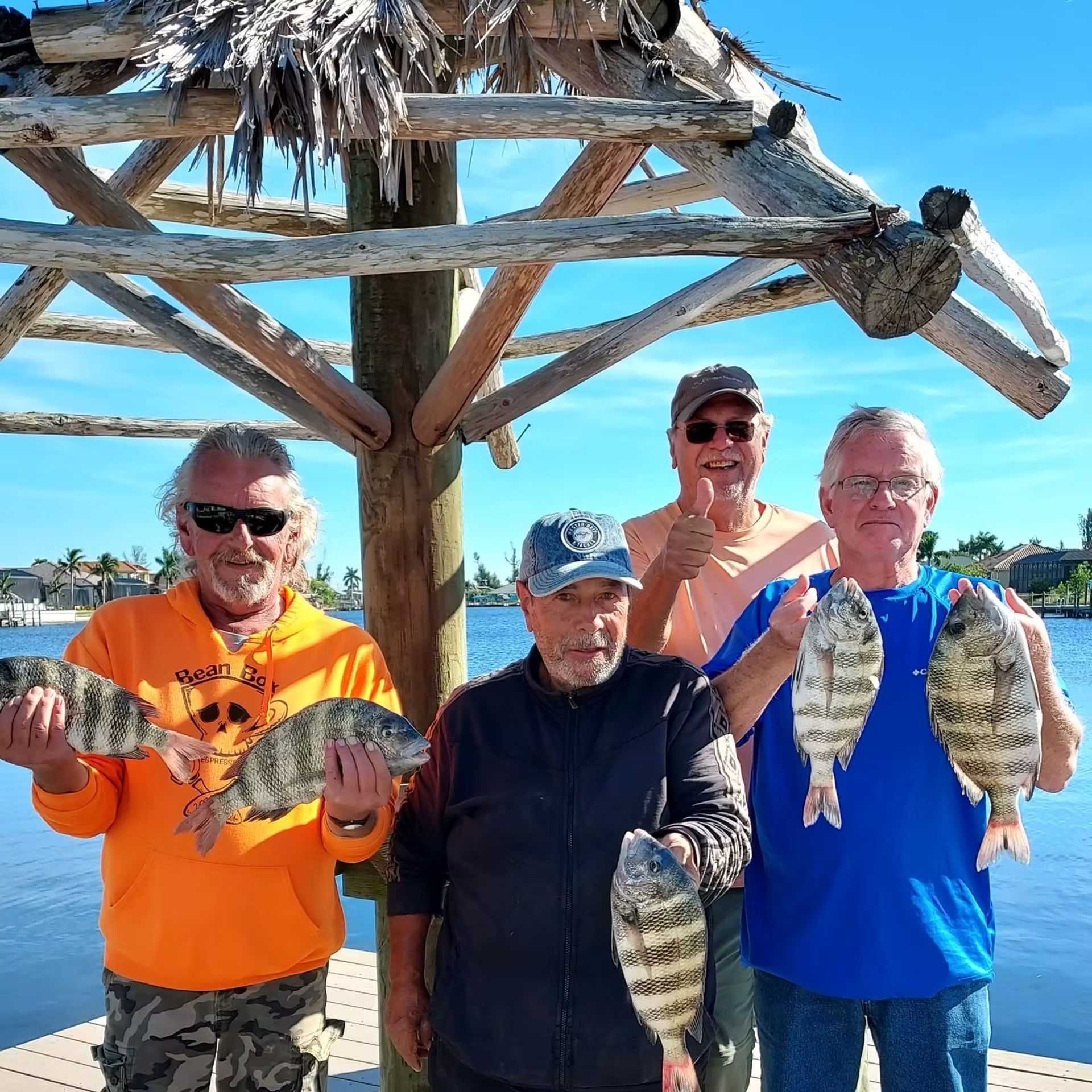 Four men pose with fish, standing by water under a tiki hut.