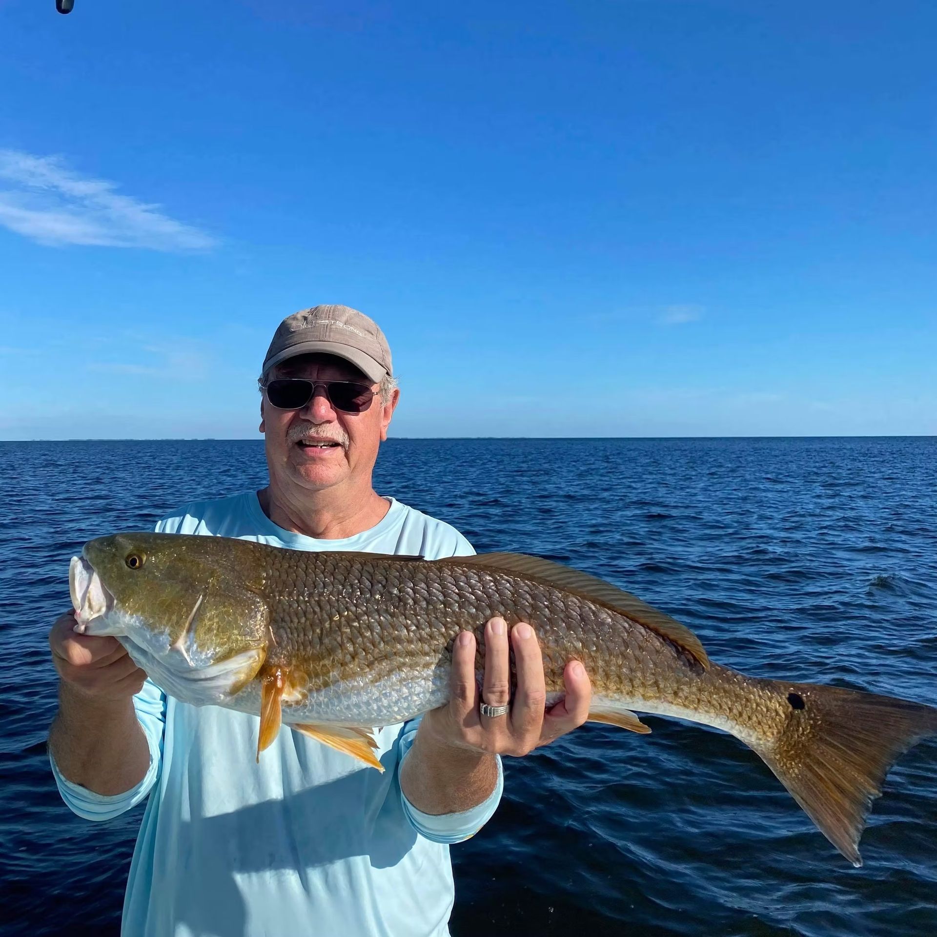 Man holding a large reddish-brown fish on a boat, with a blue sky and water background.