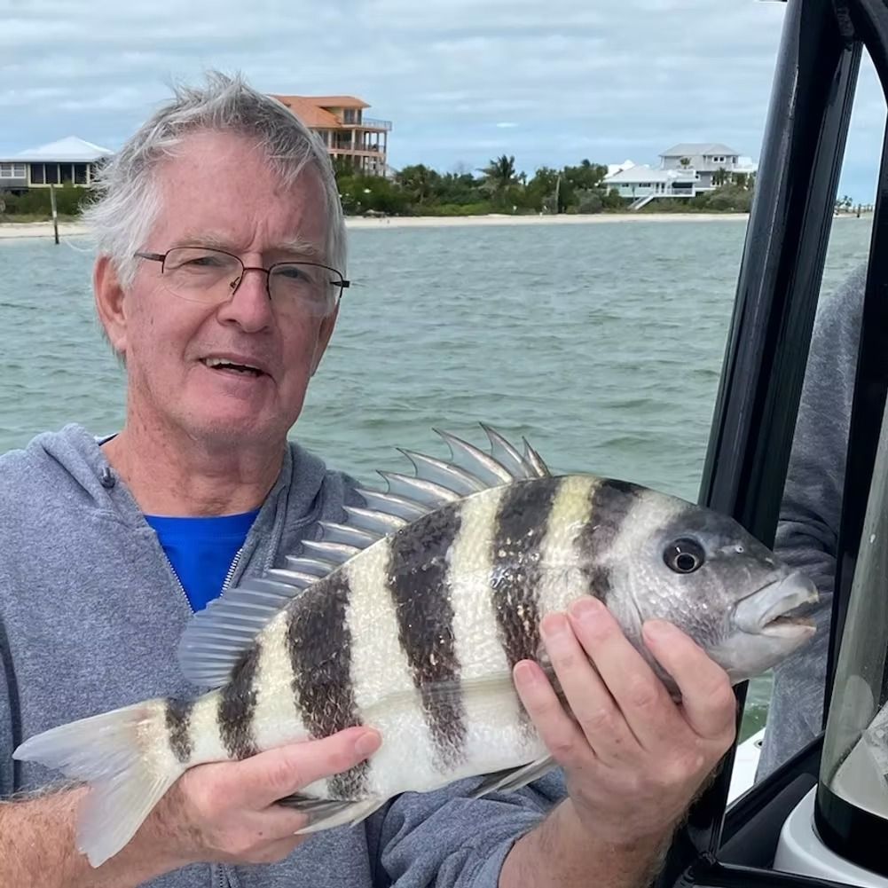 Man holding a striped fish on a boat; houses and ocean in the background.