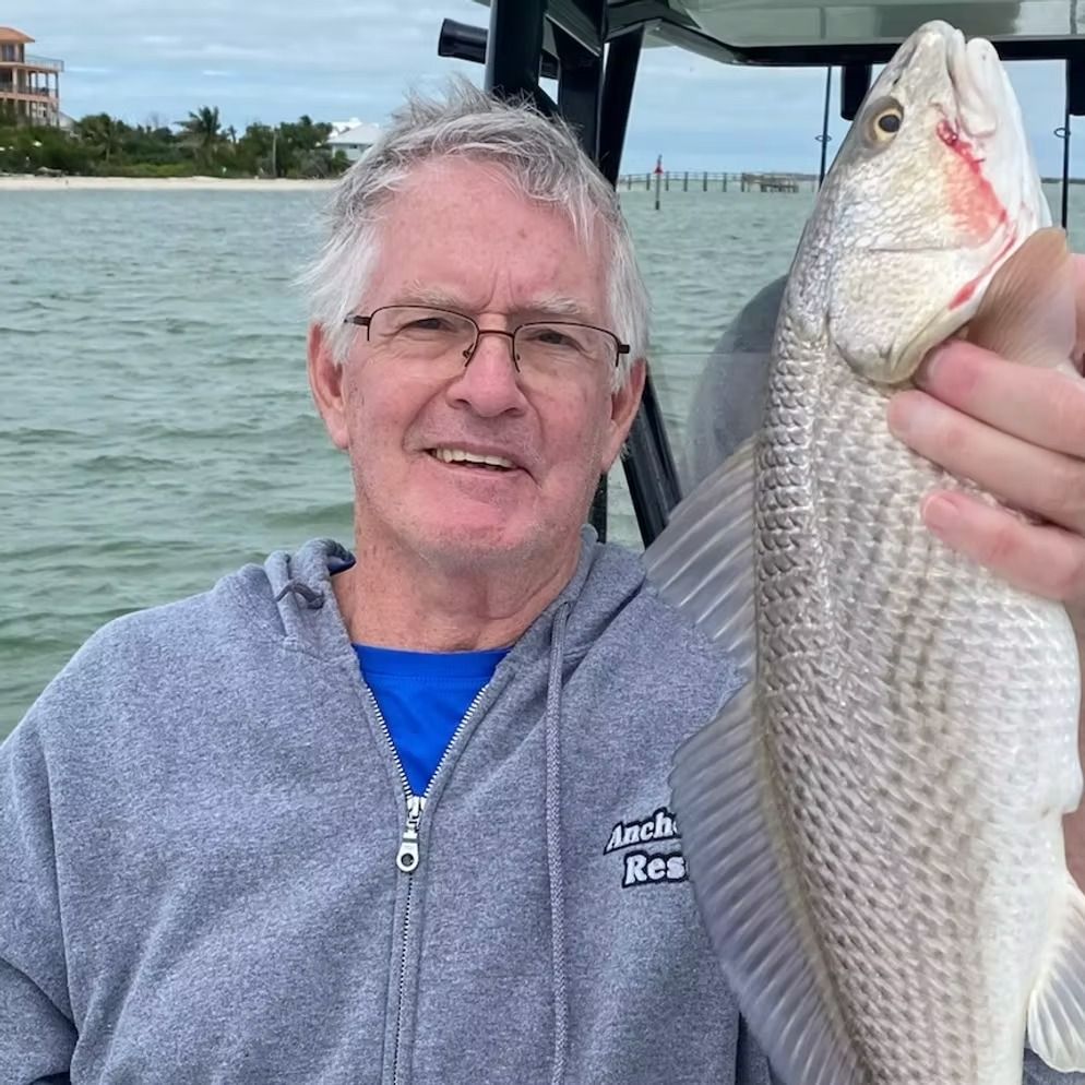 Man on a boat, smiling, holding a fish. Gray hoodie, blue shirt, water in background.