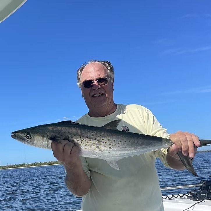 Man on boat holding a Spanish mackerel, smiling. Clear blue sky and water.