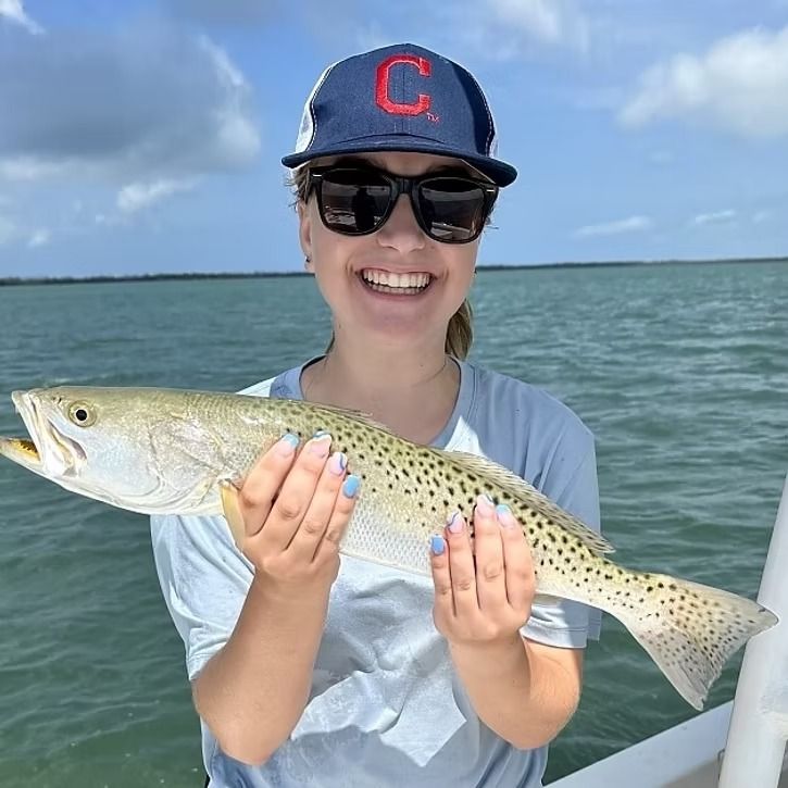 Woman smiling, holding speckled sea trout. Wearing sunglasses, baseball cap, on boat.