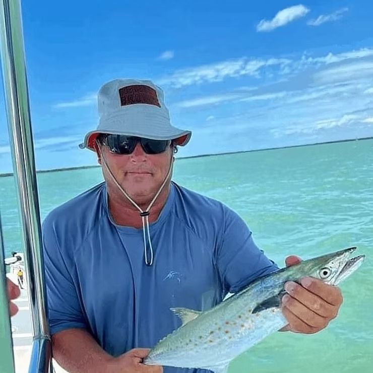 Man in hat and sunglasses holds a fish on a boat, ocean and sky in the background.