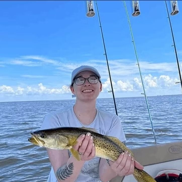 Woman smiling, holding a brown spotted fish on a boat, with fishing rods and the ocean in the background.