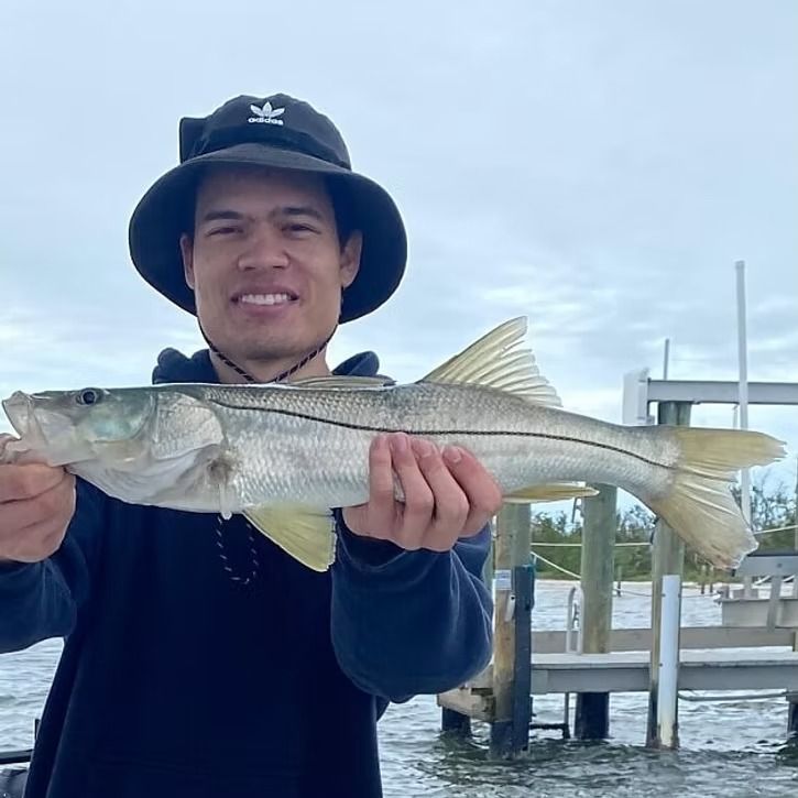 Man smiling, holding a silver snook fish next to a pier, wearing a black hat and hoodie.