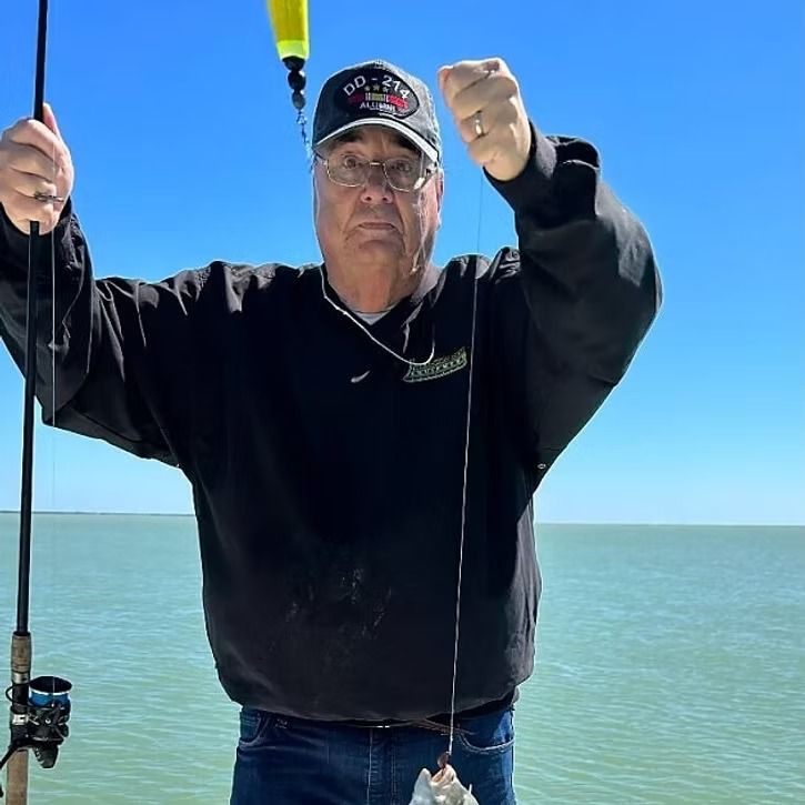 Man fishing on a pier with a blue sky, wearing a black sweatshirt and cap.