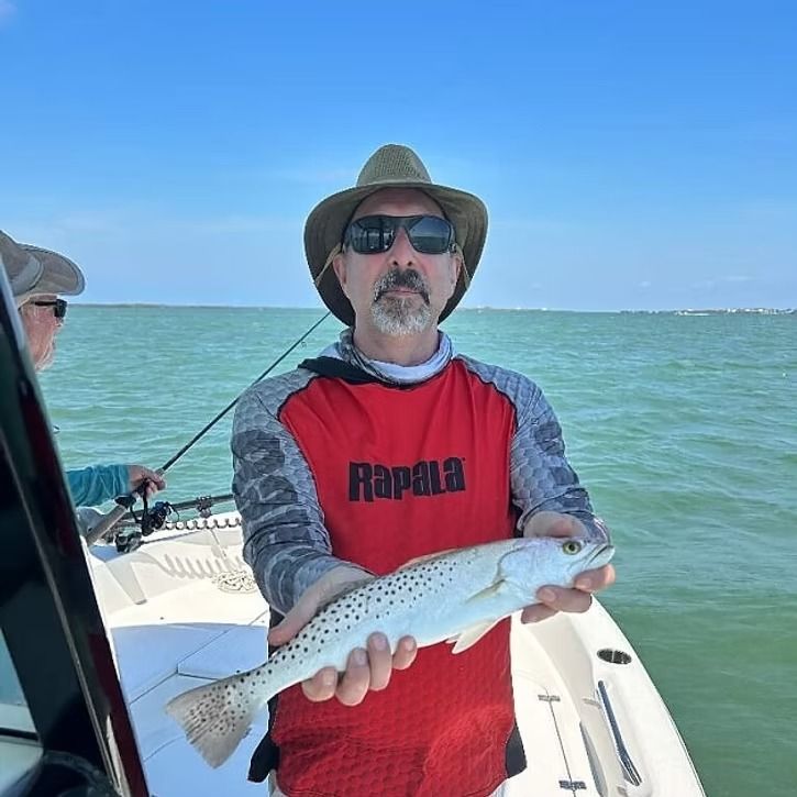 Man in hat holding speckled trout on a boat, sunny day at sea.