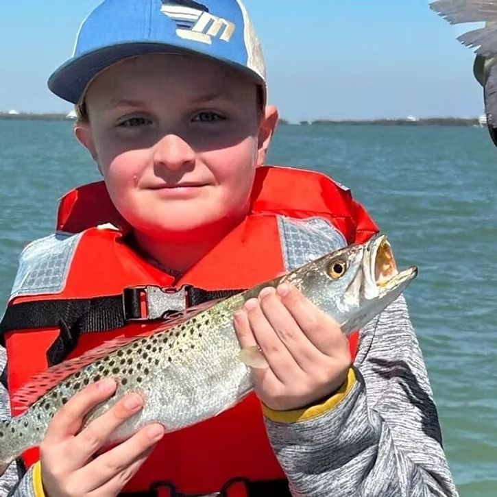 Boy in life vest holding fish, smiling on a boat, blue water background.