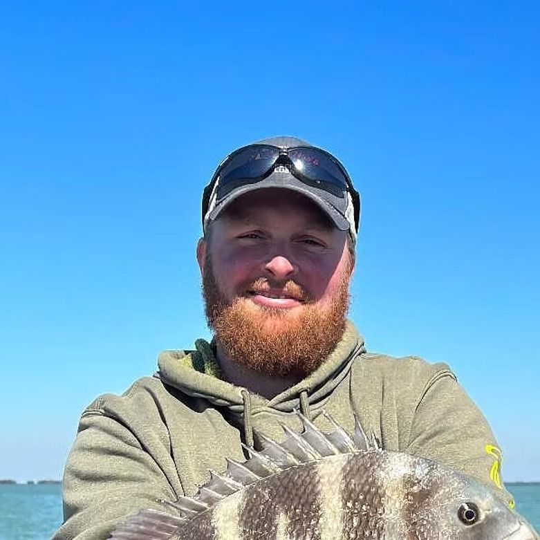 Man with red beard holding a fish, smiling, under blue sky.