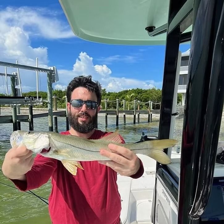 Man on boat holds up a snook fish. He wears sunglasses and a red shirt. Background includes water and dock.