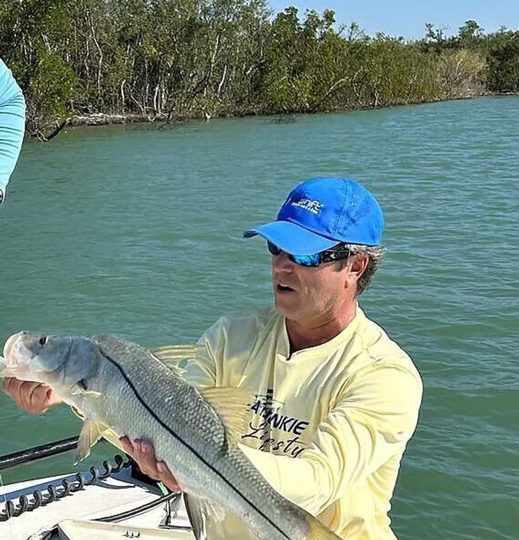 Man in blue cap holding a large fish on a boat, near mangroves.