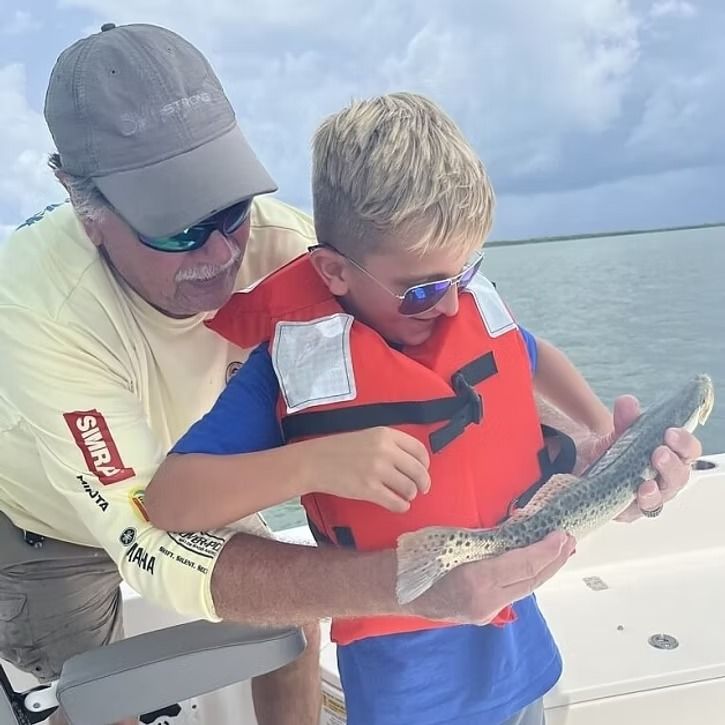 Man helps a boy hold up a speckled trout on a boat; both wear sunglasses and life vests, outdoors.