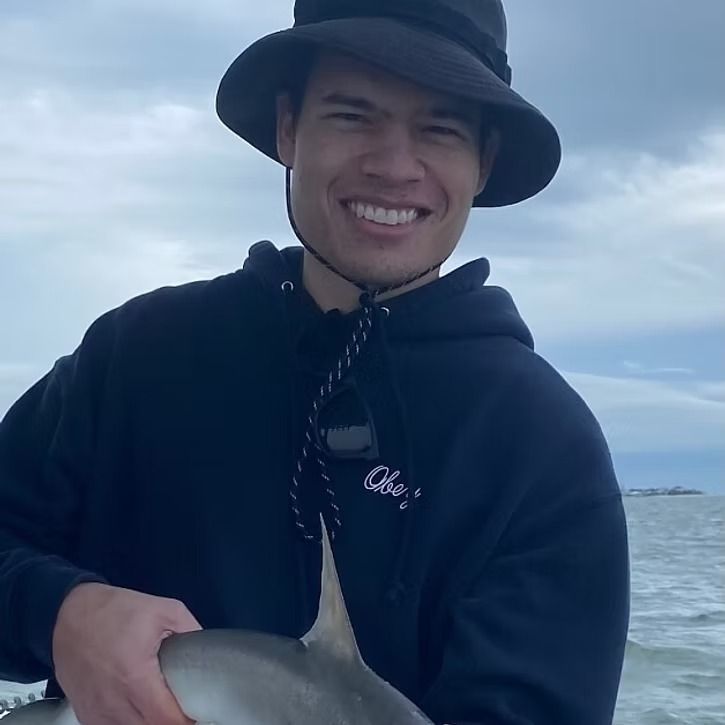 Man in hat and hoodie smiling, holding a small shark on a boat at sea.