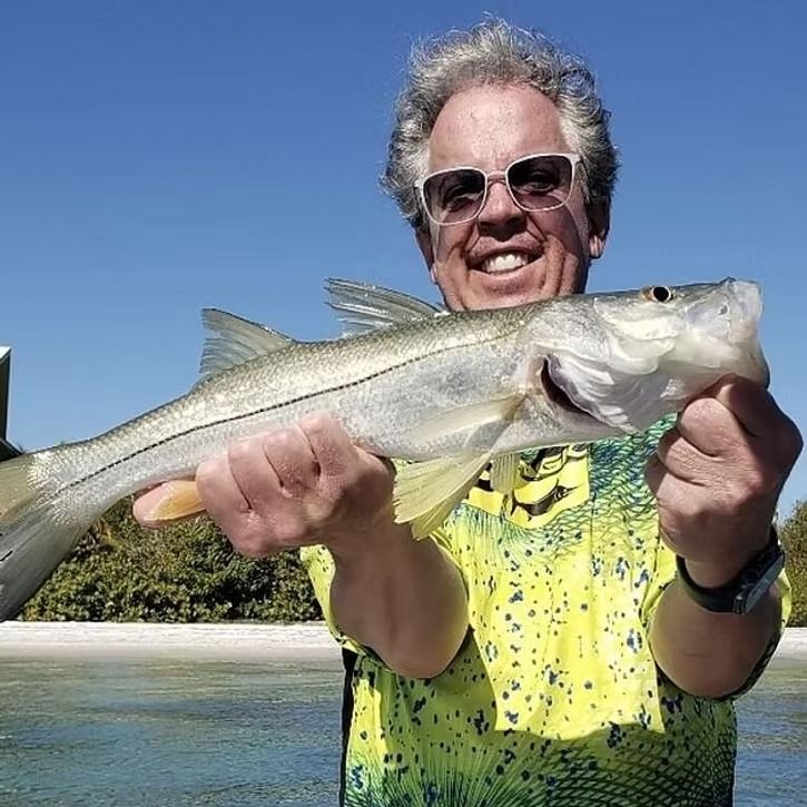 Man holding a snook fish, smiling. Blue sky, water, and beach in the background.