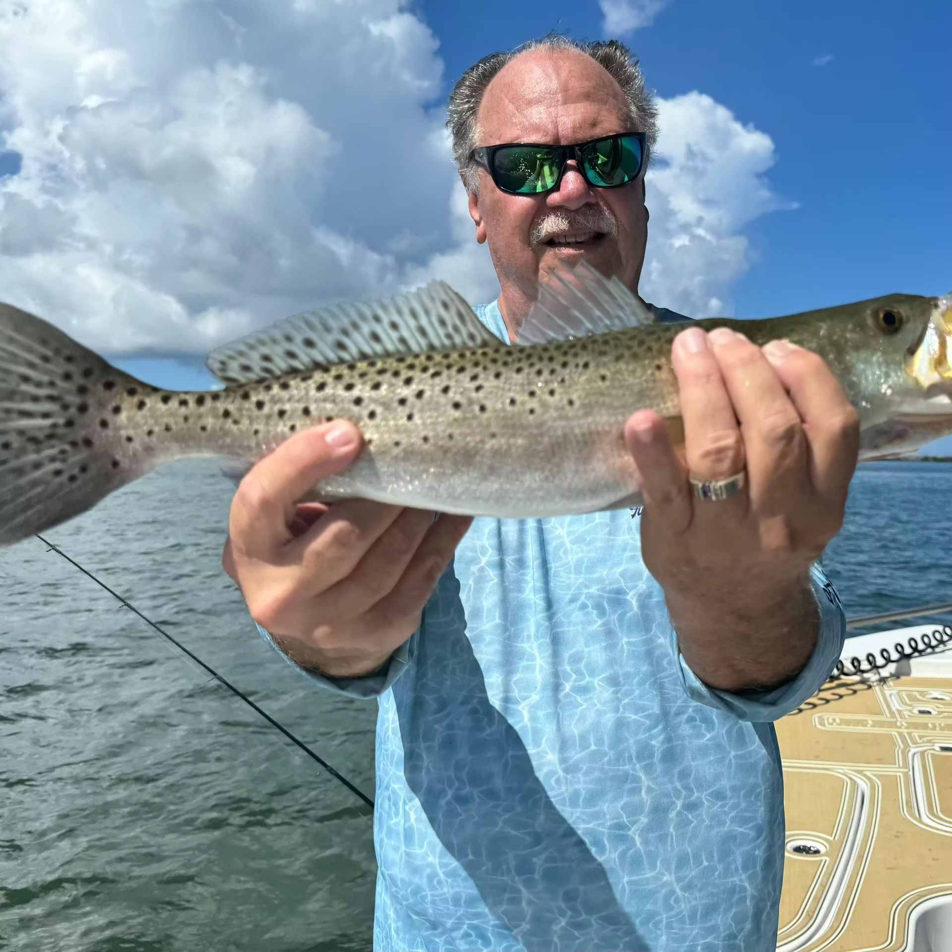 Man in sunglasses holding a speckled trout he caught on a boat, against a cloudy blue sky.