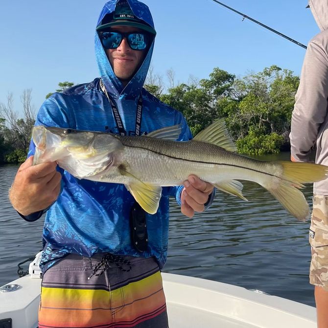 Man on a boat holding a Snook fish. He wears sunglasses, a blue hoodie, and colorful shorts.
