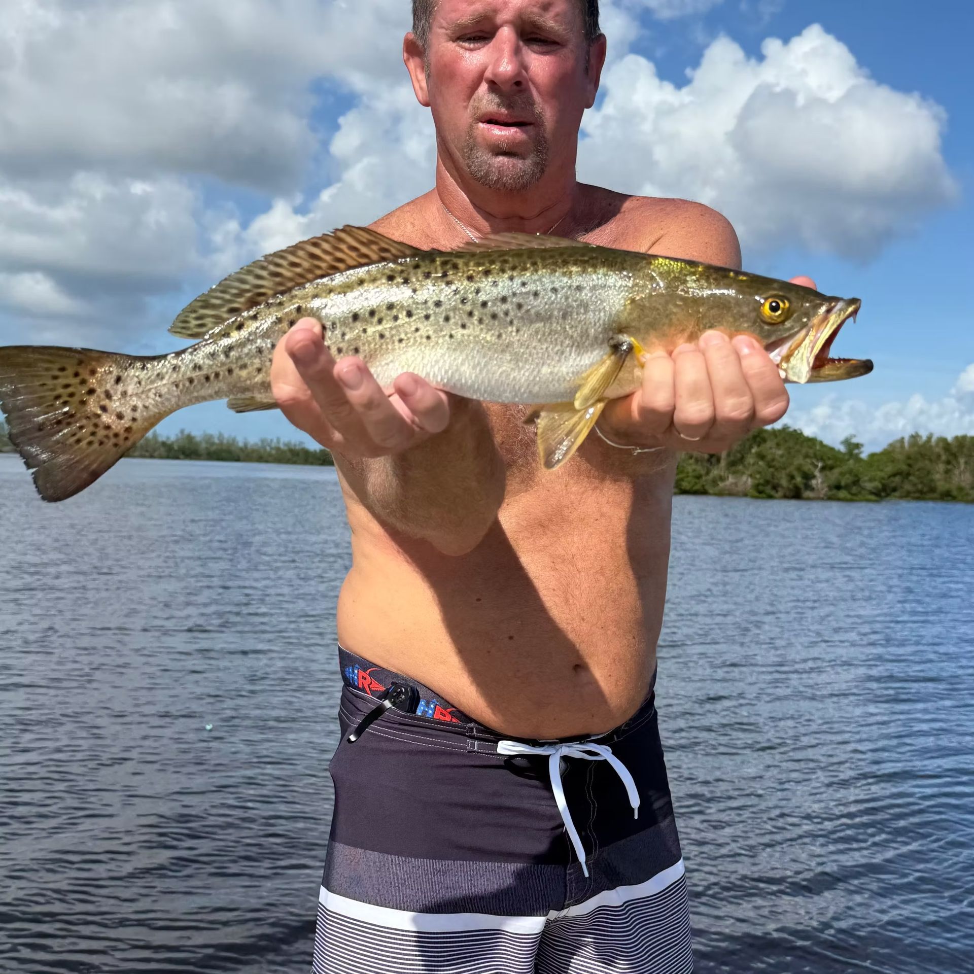 Man holding a speckled trout on a boat. Blue water, sunny sky.