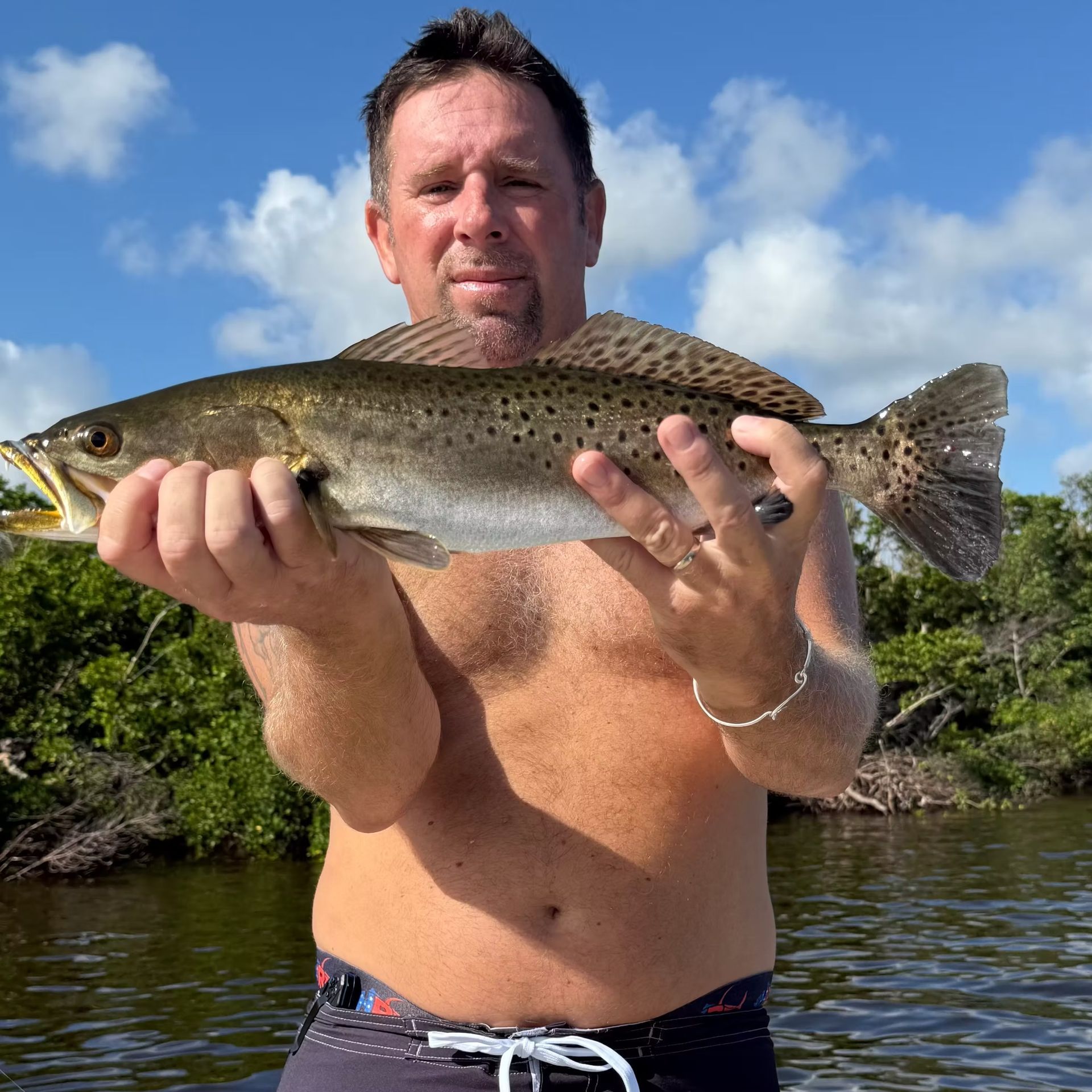 Man holding a spotted speckled trout in front of green foliage and blue sky.