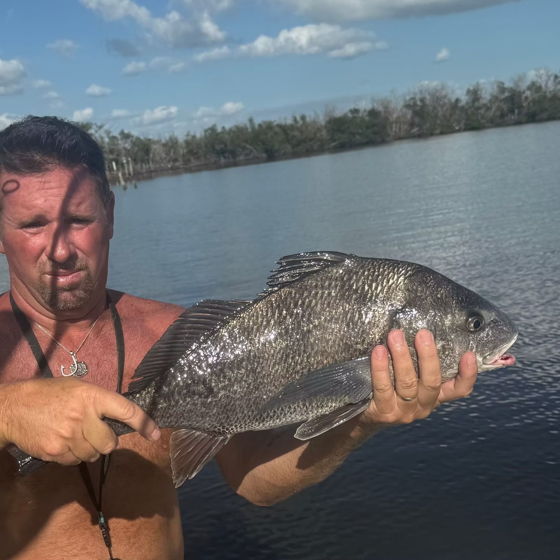 Man holding a large, dark fish. He's on a boat with water and trees in the background.