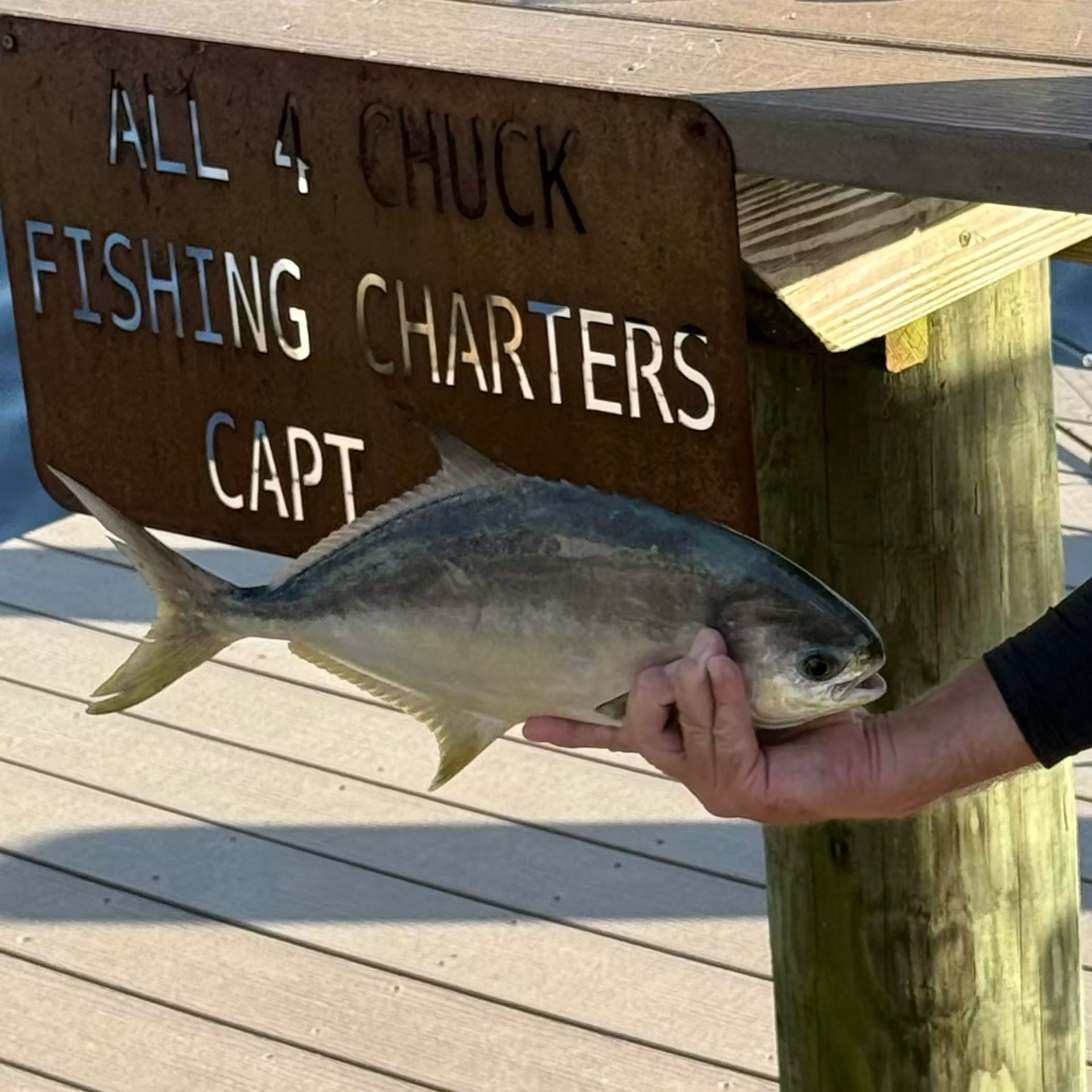 A man holding a silvery pompano fish near a dock sign for fishing charters.