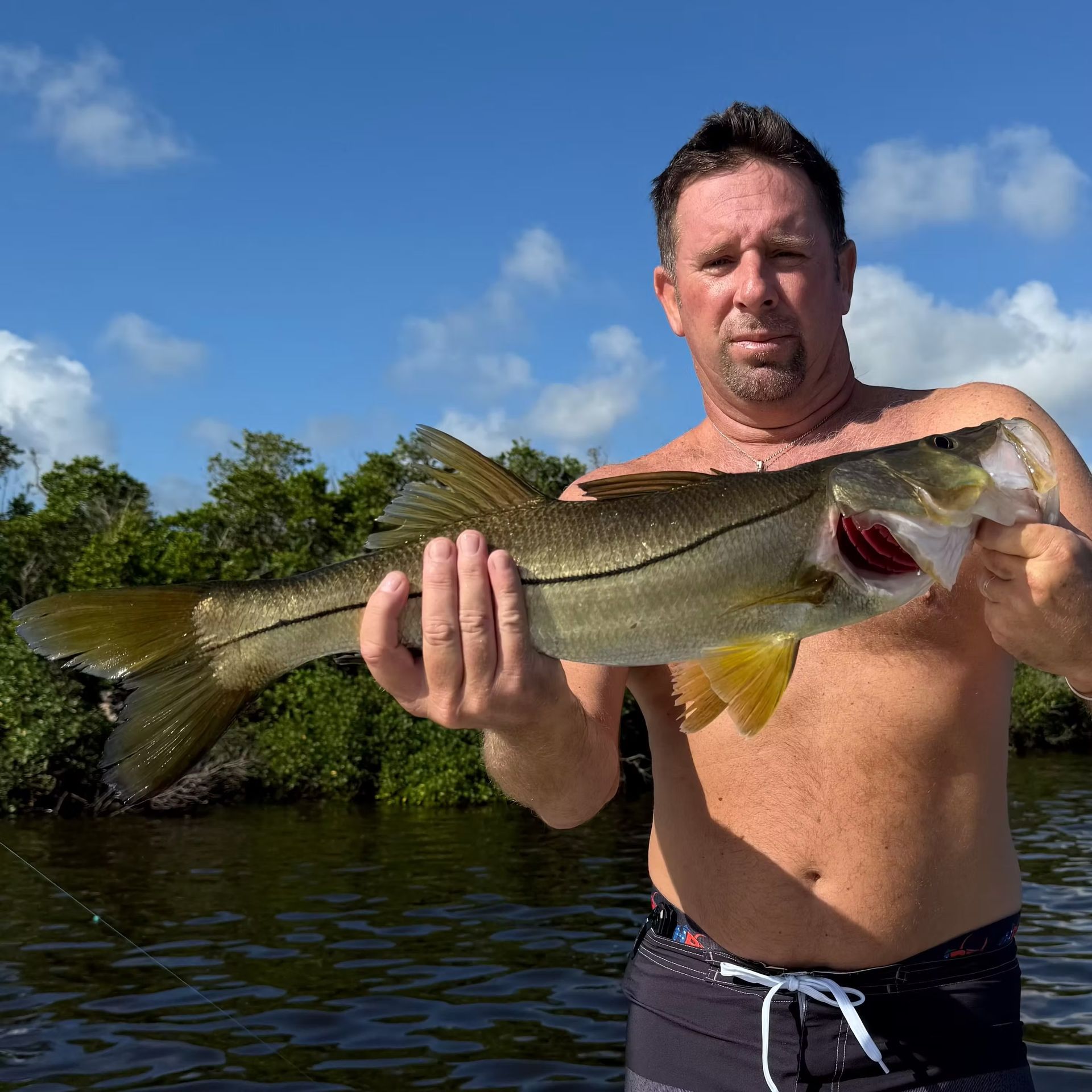 Man holding a silver snook fish on a boat. Blue sky, green trees in background.