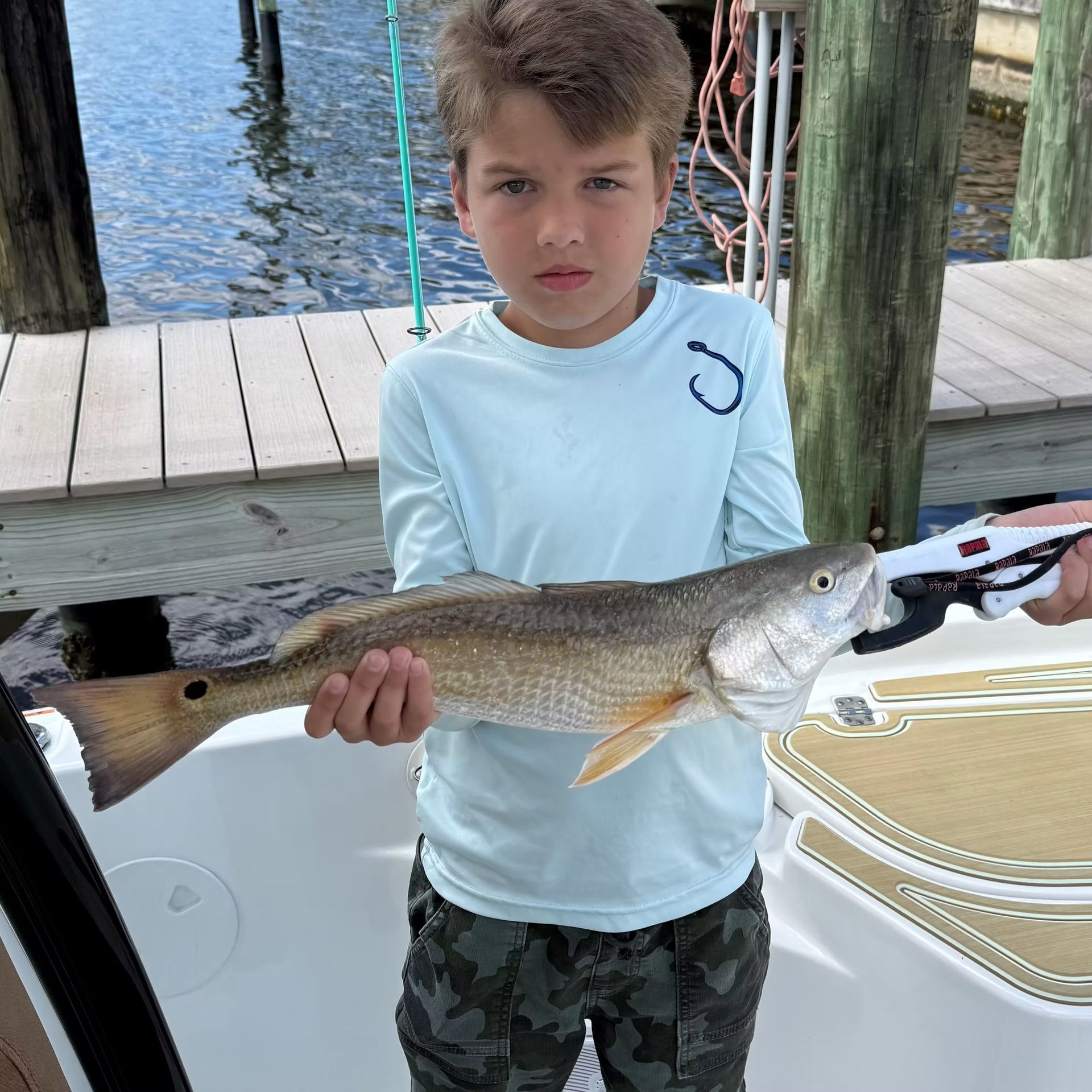 Boy holding a redfish on a boat; wearing light blue shirt, camo pants, stern look.