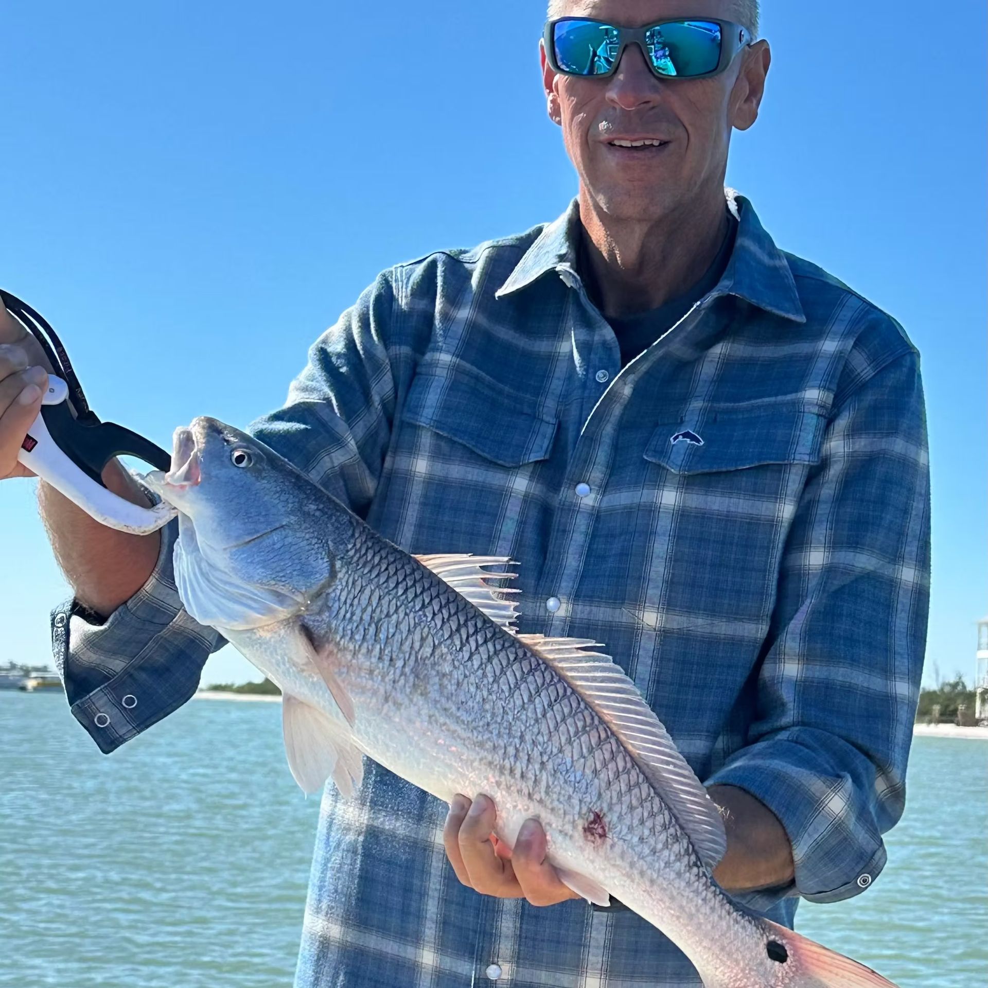 Man in sunglasses holds a Redfish on a boat. Ocean and blue sky in background.