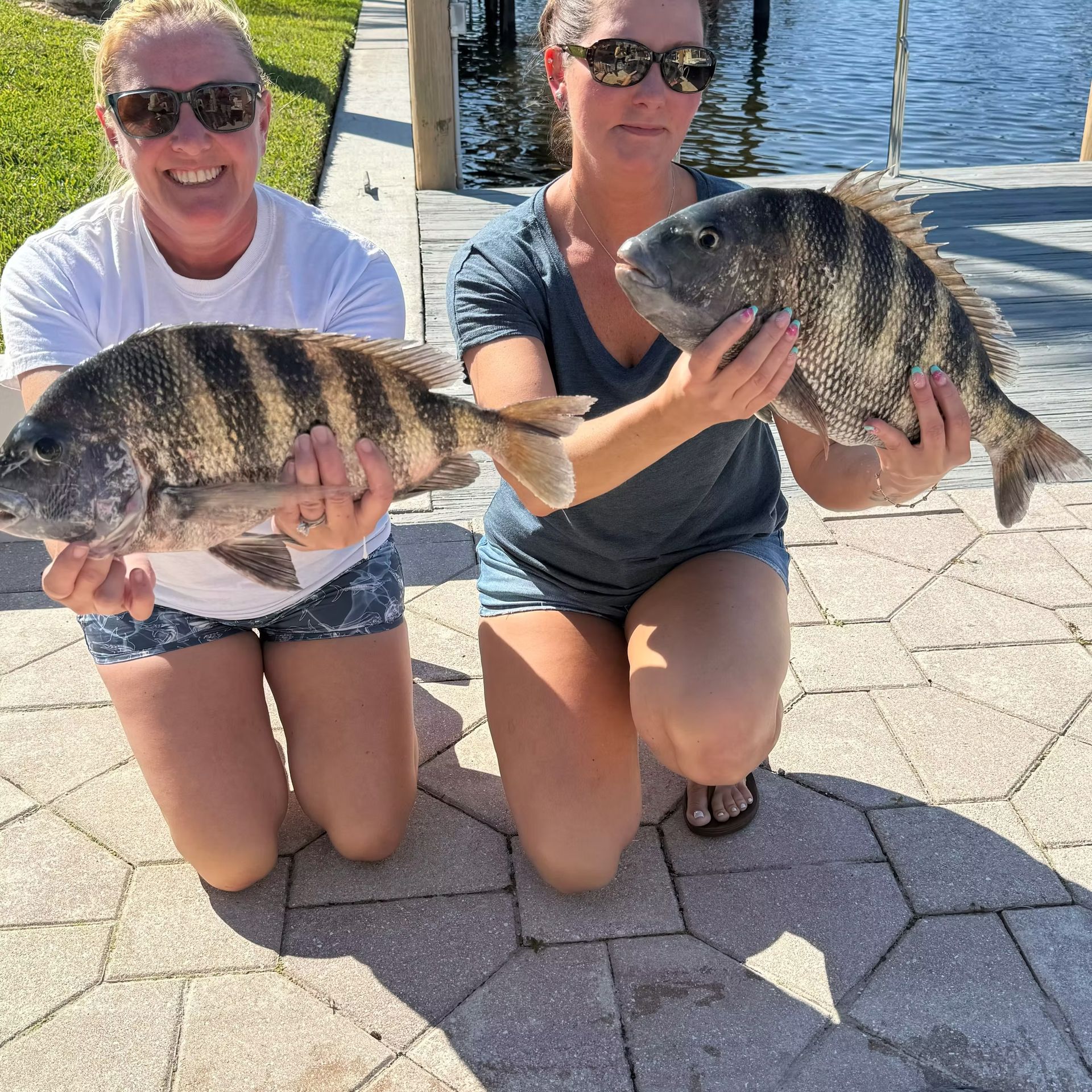 Two women kneeling, holding large striped fish near a dock.