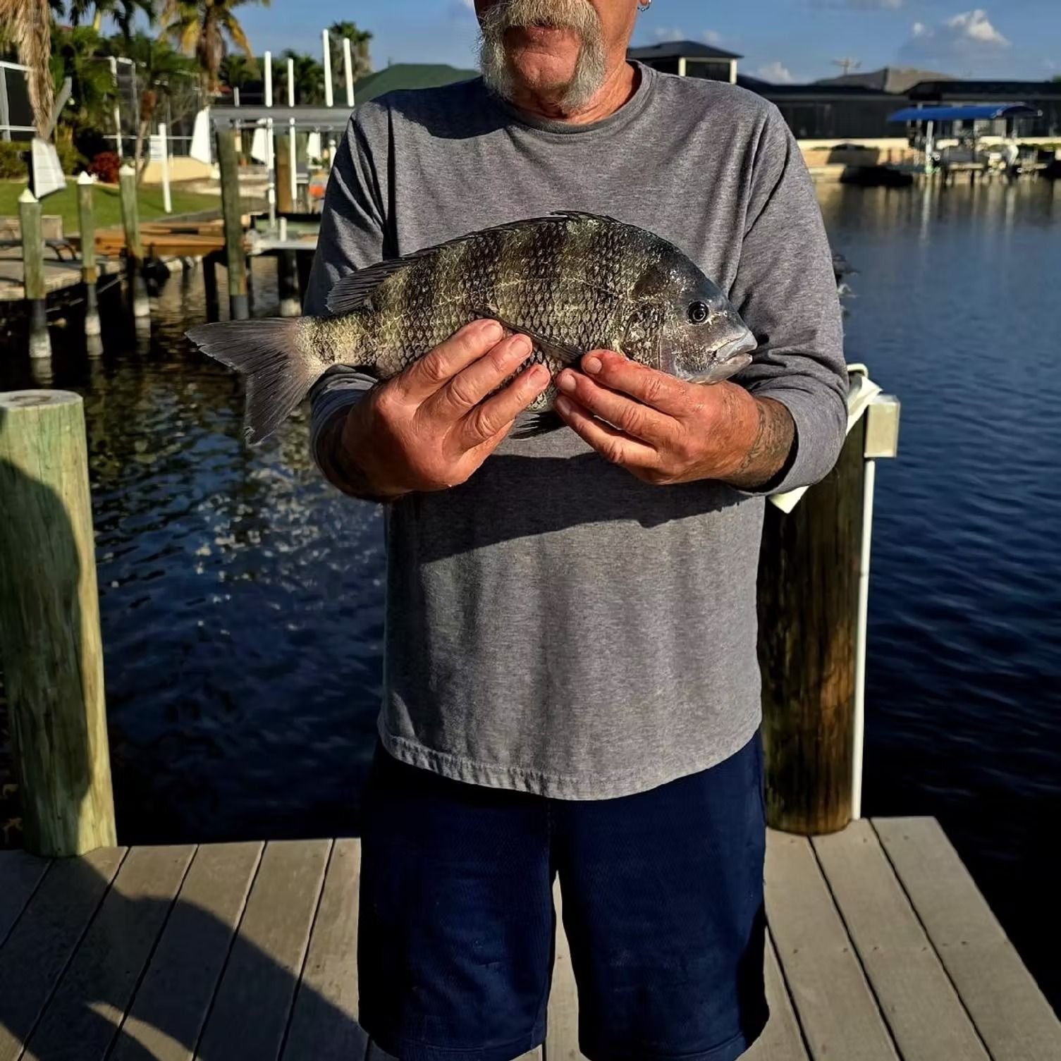 Man holding a dark-colored fish on a dock with water and houses in the background.