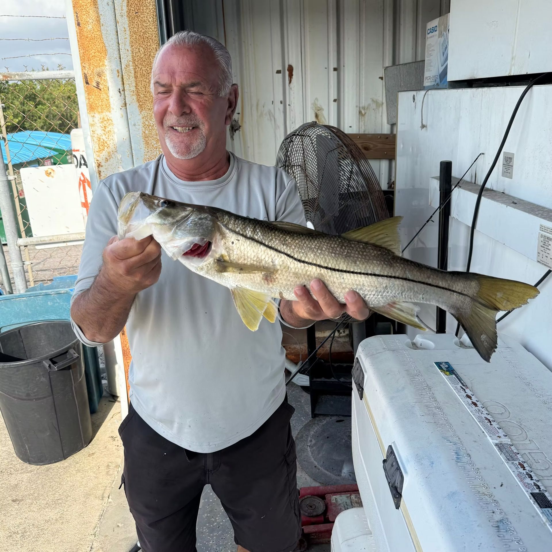 Man holding a fish, smiling. Outdoors near boat, wearing a gray shirt and dark shorts.