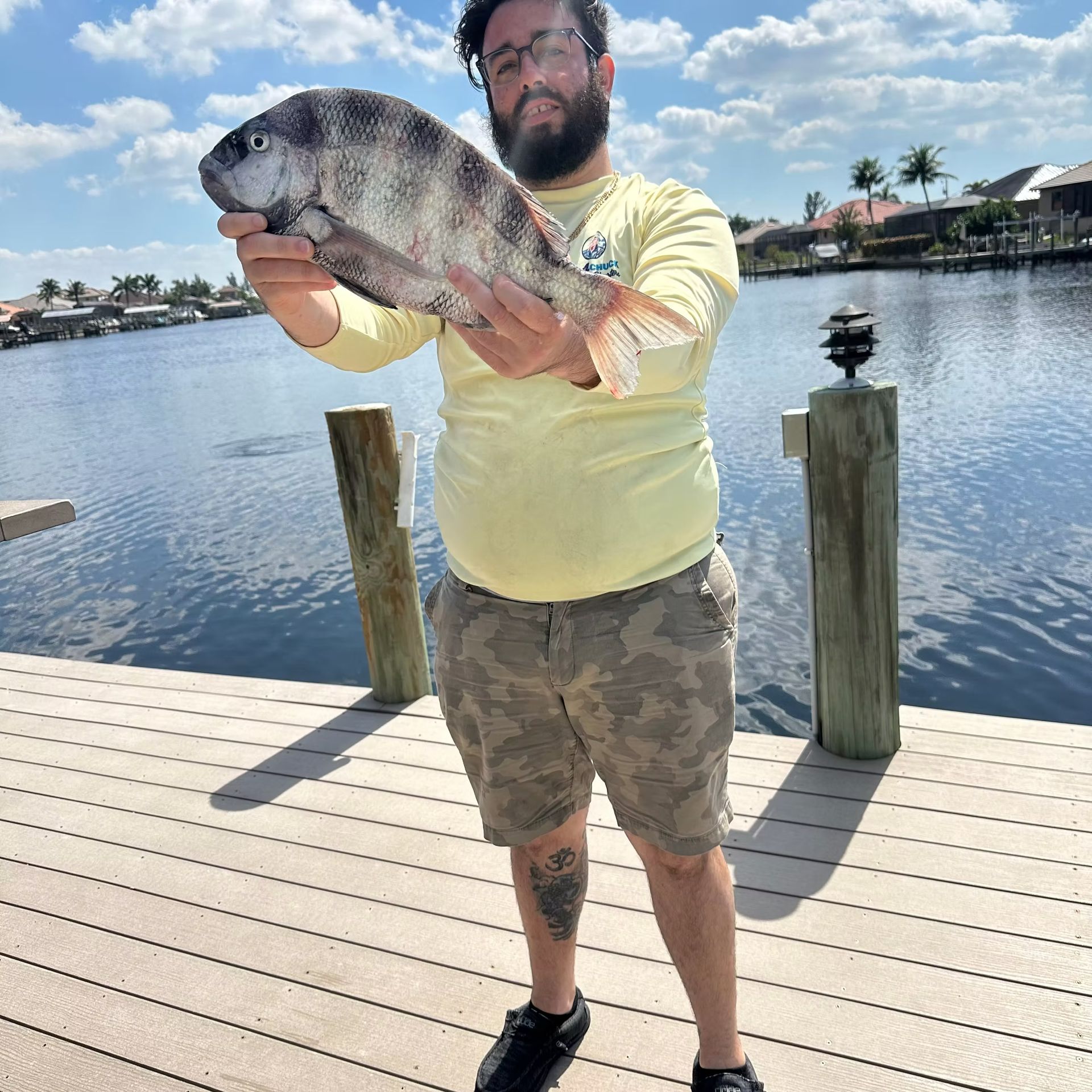 Man holding a large fish, standing on a wooden dock by a waterway on a sunny day.