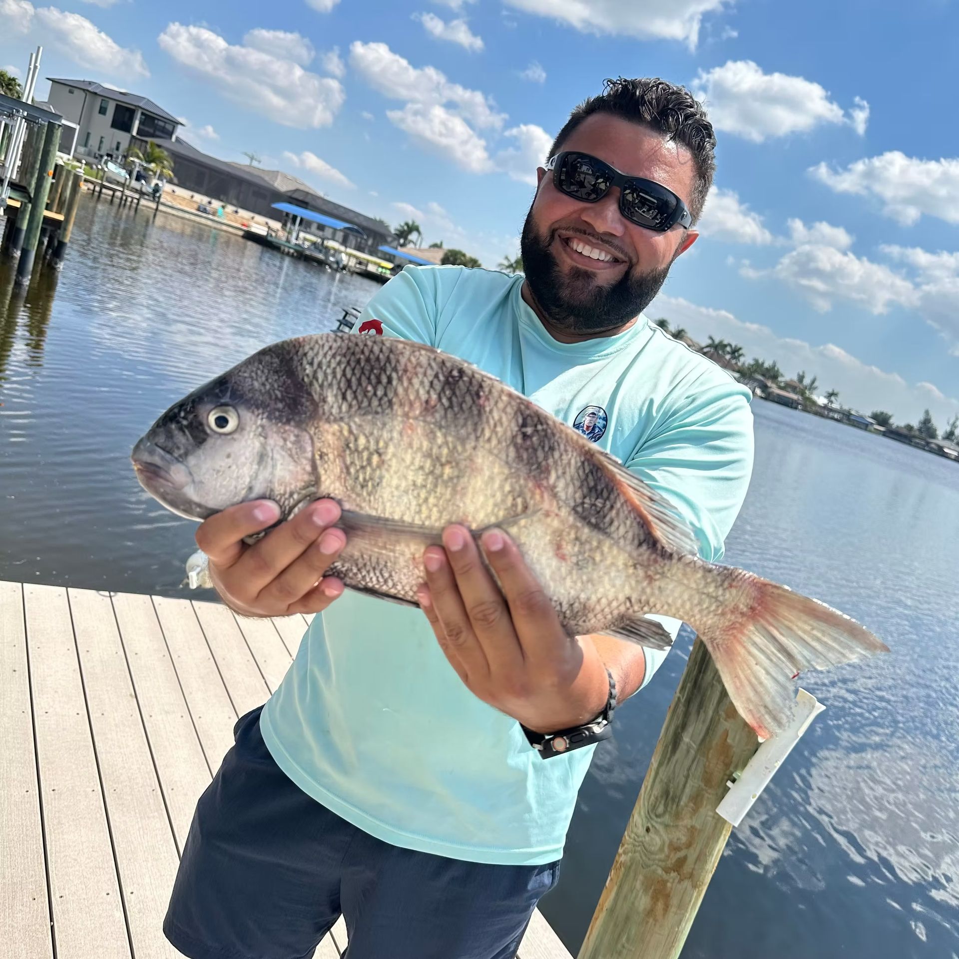 Man holding a striped fish on a dock, wearing sunglasses and smiling. Water and houses in background.
