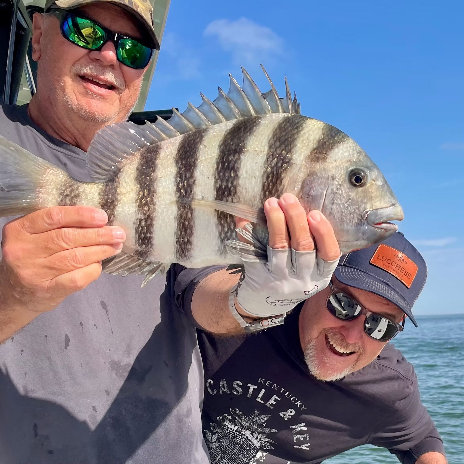 Two men on a boat, smiling while holding a striped sheepshead fish against a blue sky.