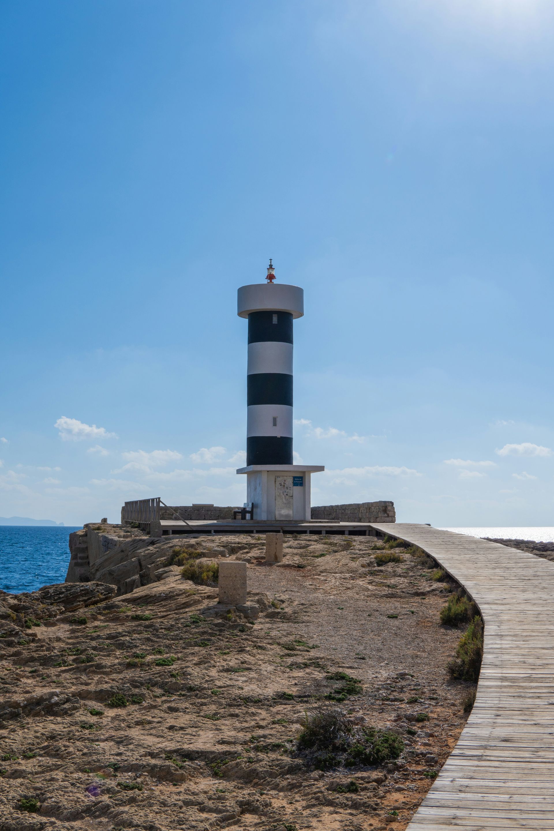 Een vuurtoren met zwart-witte strepen op een rotsachtige punt onder een helderblauwe hemel.